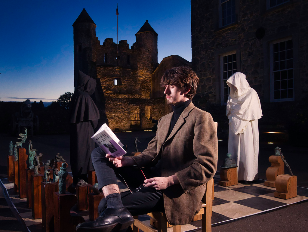 Adult man sitting on a chair reads from a book, behind two people in hoods stand on a large chessboard with wooden pieces, a castle and deep-blue sky is seen in the background.